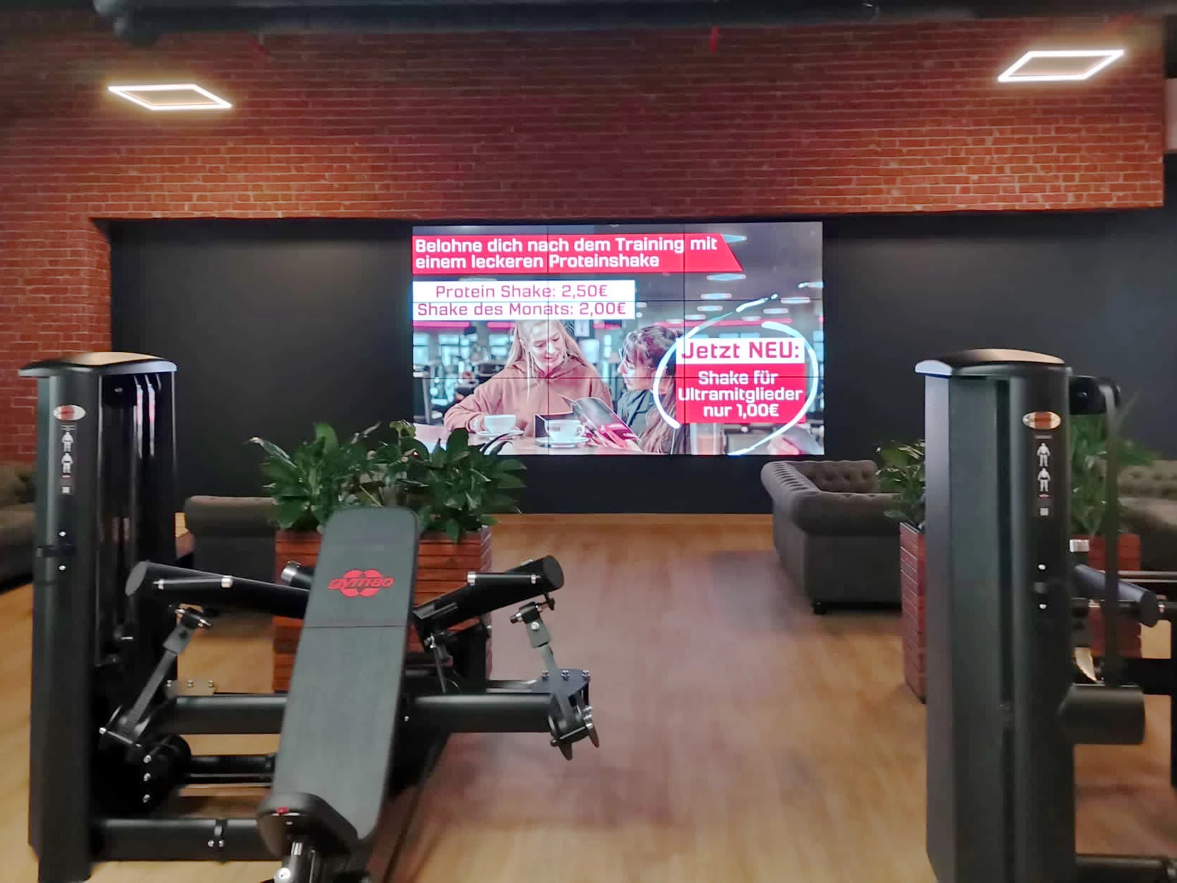 Lounge- und Trainingsbereich im Fitnessstudio mit Werbebildschirm für Proteinshake-Angebote nach dem Training