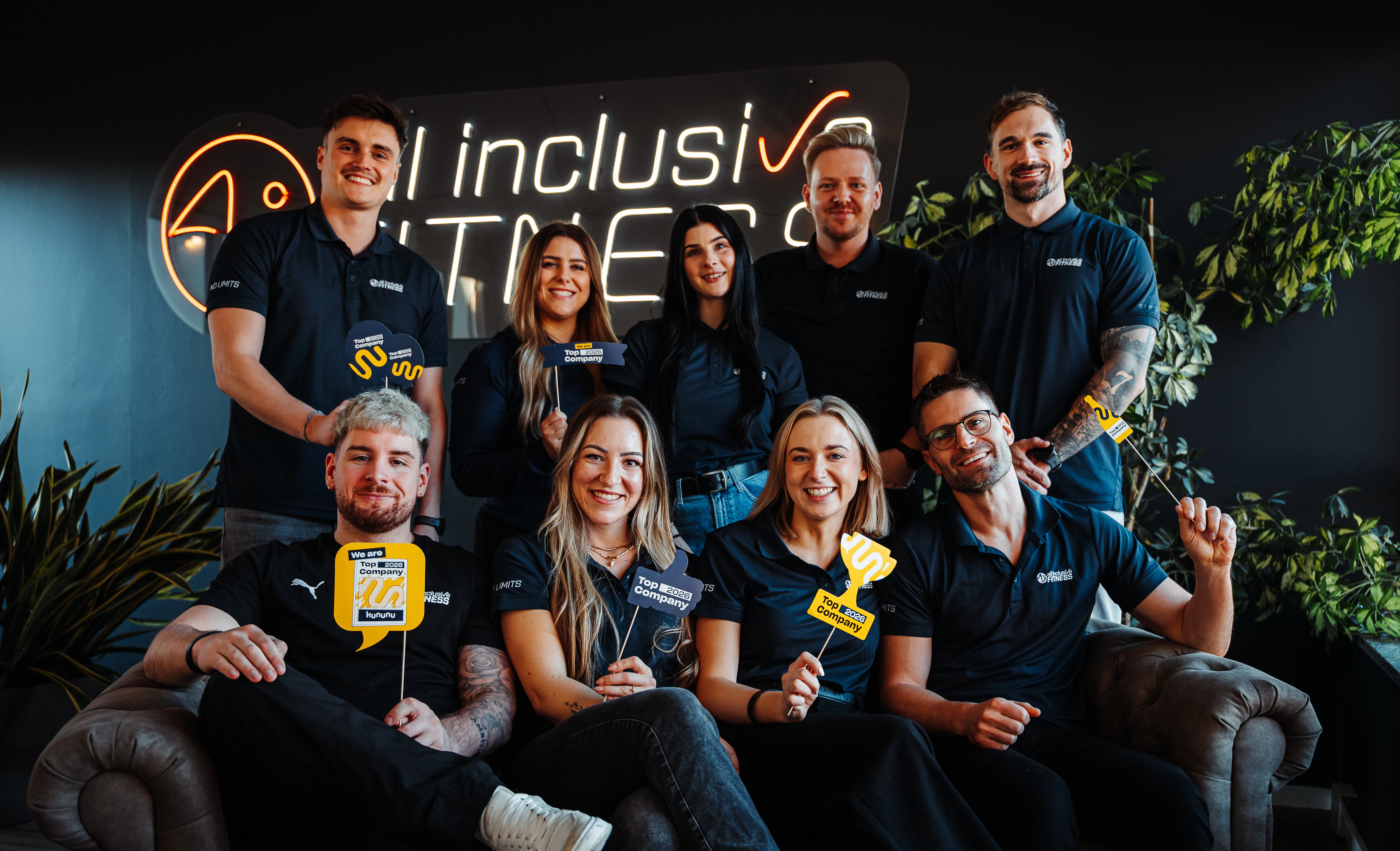 Drei Mitarbeitende im schwarzen Shirt posieren vor dem Logo von all inclusive Fitness im Studio.