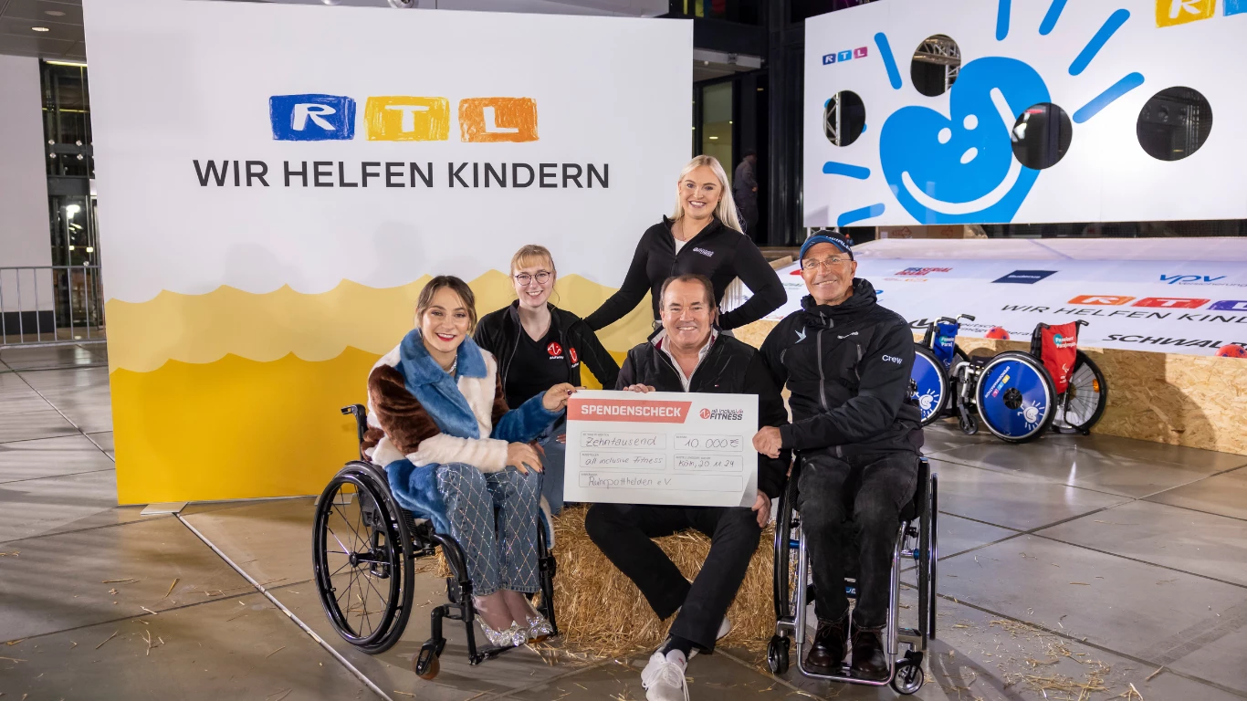 Gruppe von sechs Personen bei einer Spendenaktion für RTL Wir helfen Kindern, zwei davon im Rollstuhl, gemeinsam mit großem Spendenscheck.