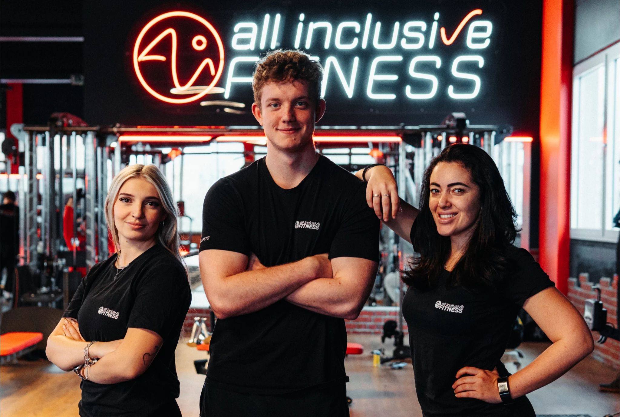 Drei Teammitglieder von all inclusive Fitness stehen nebeneinander im Studio vor dem Logo. Sie tragen schwarze Shirts mit dem Studio-Logo und lächeln selbstbewusst in die Kamera