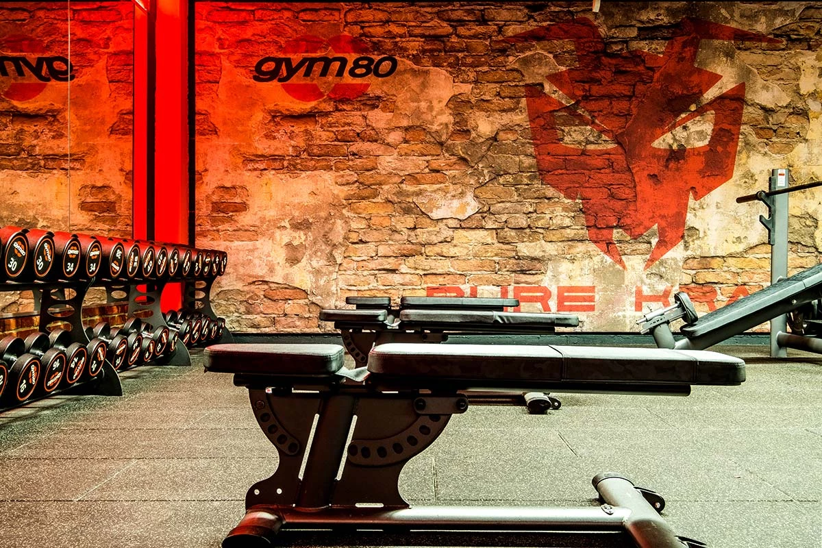 Freihantelbereich mit Hantelbänken und Kurzhanteln vor rustikaler Backsteinwand mit gym80-Branding