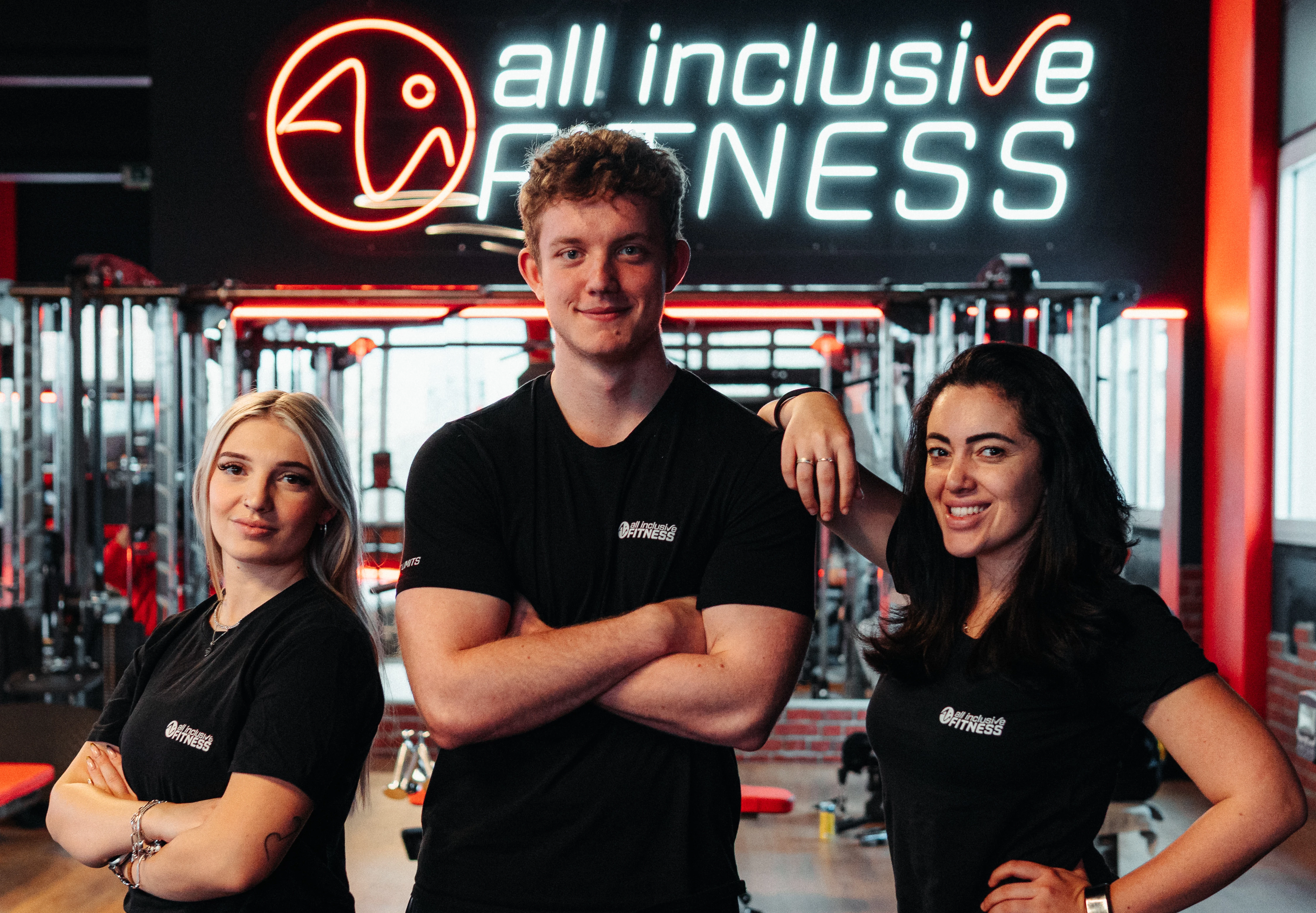 Drei Mitarbeitende im schwarzen Shirt posieren vor dem Logo von all inclusive Fitness im Studio.