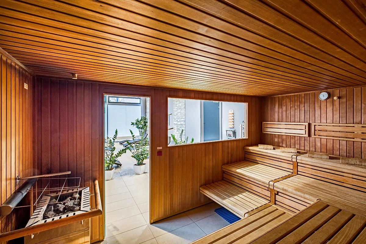 Holzsauna im Fitnessstudio mit mehreren Sitzbänken, Saunaofen links und Blick durch eine Glastür in den Ruheraum mit Pflanzen