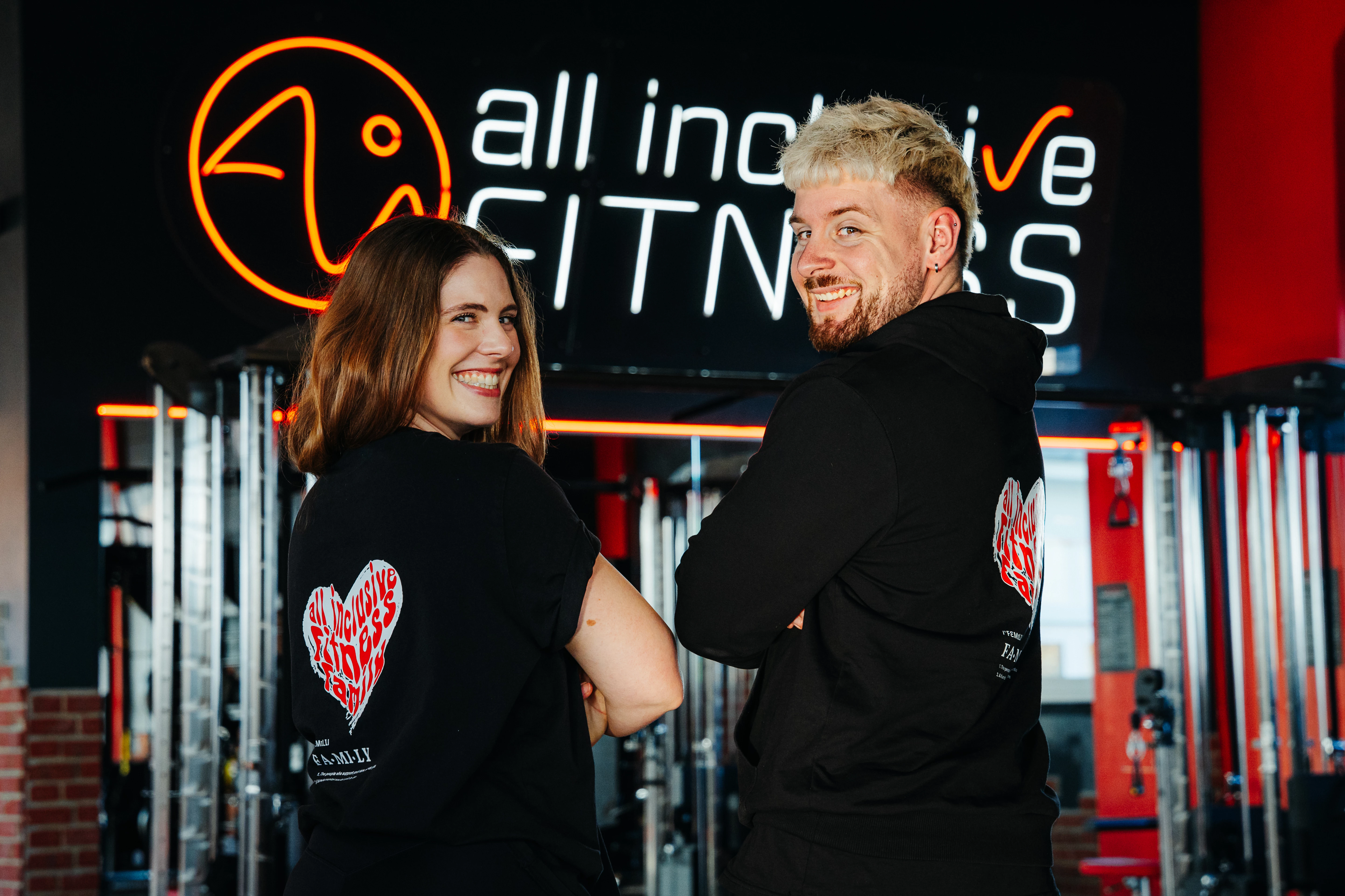 Rückansicht eines Mitarbeiters im roten Shirt mit der Aufschrift TEAM, der mit drei Personen am Tresen im Fitnessstudio spricht