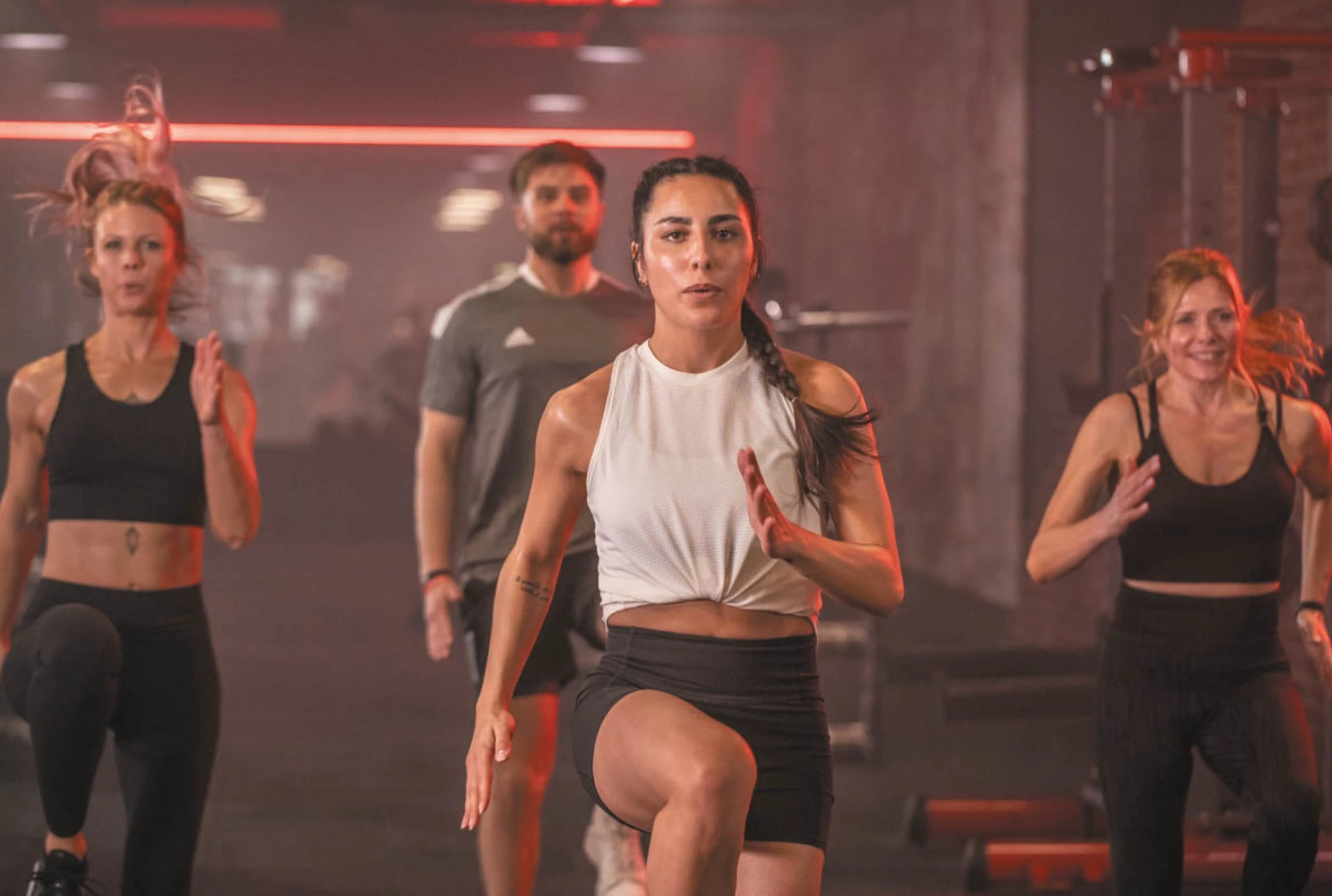 Mehrere Personen trainieren gemeinsam in einem Fitnessstudio. Im Vordergrund führt eine Frau in weißem Sporttop eine dynamische Übung an, während weitere Teilnehmerinnen und ein Teilnehmer im Hintergrund motiviert mitmachen.