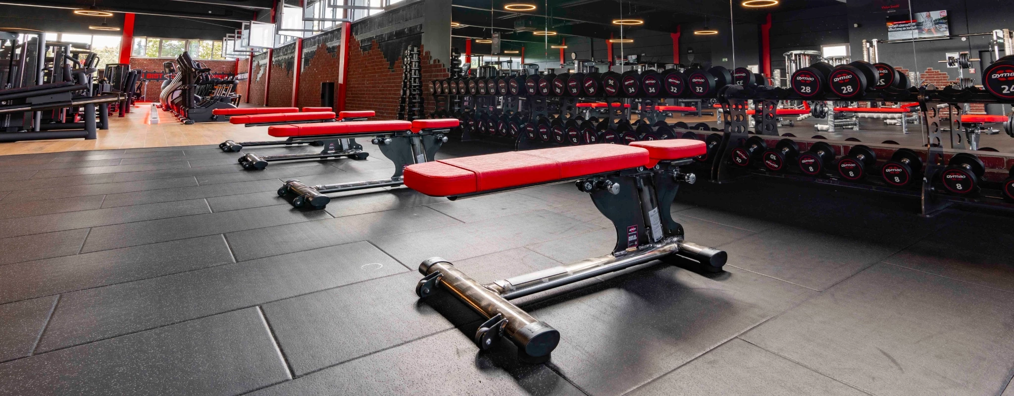 Hantelbereich im Fitnessstudio mit langen Reihen an Kurzhanteln und roten Trainingsbänken, beleuchtet von rotem Licht