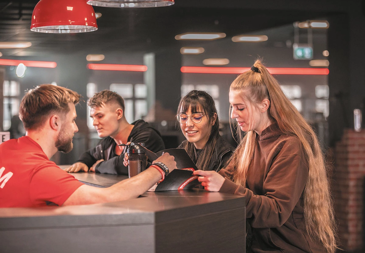 Mitarbeiter im roten Shirt spricht mit drei Personen am Tresen im Fitnessstudio.