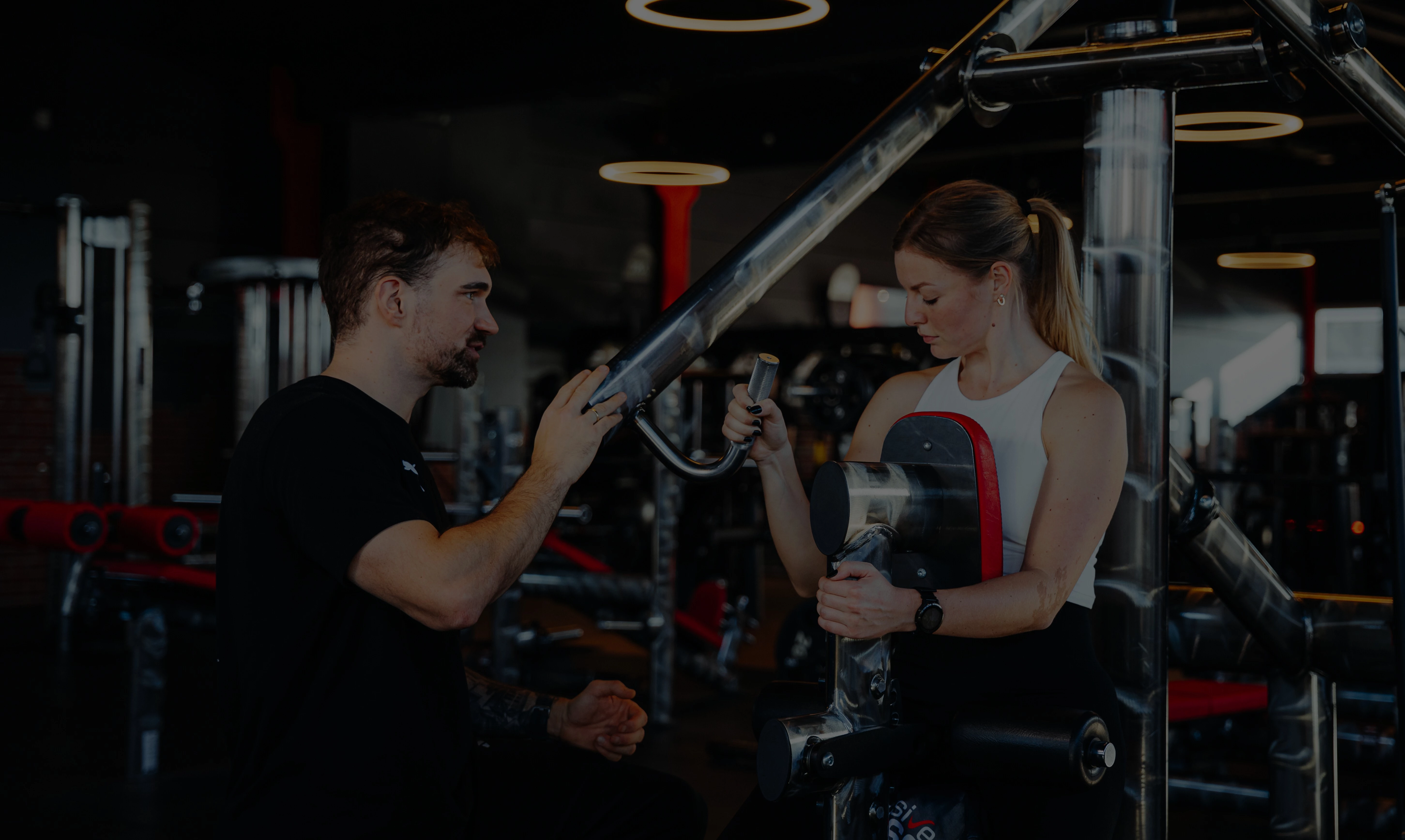Zwei Personen auf einem Luftbike geben sich die Faust. Im Hintergrund ist ein ausgestattetes Fitnessstudio zu sehen.